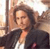 A pixelated image shows a person with long brown hair wearing a dark jacket over a white shirt, seated with one arm raised against a light-colored background.