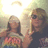 Two people pose together; one wears a cowboy hat and pink floral top, the other dark clothing with sunglasses. Bright light source behind them creates halo effect.