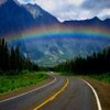 A winding road with yellow and white lines curves through a landscape featuring green grass, dark trees, mountains, and a rainbow under a cloudy sky.
