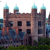 Pixelated image of a multi-story stone building with four towers, each capped with light blue spires, set against a bright sky with greenery below.