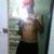 A shirtless person with short pink hair stands in a doorway holding a camera, wearing dark pants and positioned against a light-colored wall.