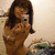 A person with long brown hair takes a mirror selfie with a silver camera, wearing a brown top, in a room with a cluttered background.