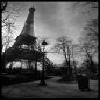 Black-and-white pixelated image of a tall tower with a lattice structure, surrounded by trees and street lamps under a cloudy sky.