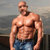 Pixelated image of a shirtless man with a beard standing outdoors near water, wearing blue jeans.