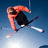 A pixelated figure in an orange jacket and black shirt performs a jump with crossed ski poles against a blue sky background.
