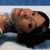 A pixelated close-up of a person lying down with eyes closed, wearing dark clothing against a blue and white background.