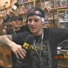 A person in a black graphic t-shirt and bandana holds a microphone with yellow logo, standing before shelves of collectibles while another person raises an object behind them.