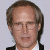A pixelated image of a person with short brown hair and glasses, wearing a dark jacket over a white shirt against a light gray background.