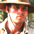 Pixelated image of a person wearing a wide-brimmed hat and light-colored shirt, with blurred green foliage in the background.