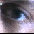 A pixelated close-up of a human eye with a blue iris and visible reflection, set against a blurred background of muted grays and whites.