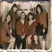 Static group portrait of seven people in brown and green clothing against a light background with visible pixelation.