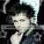 Static pixelated image of a person with dark hair and light skin wearing a dark jacket over a light shirt, set against a dark background with light and dark pixel clusters.
