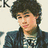 Pixelated image of a person with curly hair wearing a dark jacket over a patterned shirt, accompanied by green pixelated text on black background.