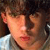 Pixelated close-up of a person's face with brown hair and light skin, wearing a white shirt against a dark background.