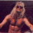 Pixelated static image of a person with long blonde hair, dark sunglasses, and muscular build against dark background.