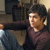 A person with curly dark hair wearing a maroon long-sleeve shirt and blue jeans sits with one arm extended, leaning against a wall in a room with wooden flooring and dark furniture.