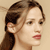 Pixelated close-up of a person with brown hair tied back, light skin, and red lips against a beige background.