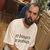 A pixelated static image shows a person in a white shirt with dark text on the chest, standing against a blurred background with brown and green tones.