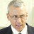 Pixelated portrait of a person with short gray hair, wearing glasses, a dark suit jacket, and a white shirt against a light background.