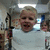 A child in a white shirt smiles widely with mouth open, standing indoors under fluorescent lights.