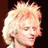 Pixelated close-up of a person with light-colored hair styled in an upward-sweeping manner against a dark background.
