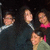 Group of people in dark clothing with one person in pink top, some faces blurred, against dark background.