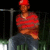 Pixelated figure in red cap and red striped shirt stands against dark background with metal railing and greenery.