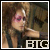 Pixelated image of a person with light brown hair and pink eyeshadow, wearing dark clothing, extending an arm forward against a blurred background.