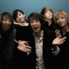 Five people pose closely together against a solid blue background, with one person in the center wearing a white shirt under a black jacket.