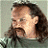 Pixelated static image of a person with long dark hair and a beard, wearing a dark shirt against a muted greenish background.
