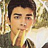 A person with short dark hair holds two yellow french fries in their mouth against a blurred background of trees and light.