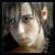 Pixelated static image of a person's face with light skin tone, dark hair, and visible ear against a blurred background.