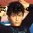 A person with dark hair in a black shirt with blue accents, hands behind head, against a red and blue background with partial text visible.