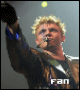 A person with curly blonde hair wearing a dark jacket with blue accents and a microphone, raising one arm against a gray background with a yellow light, with the word 'fan' at the bottom.