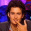 A pixelated image of a person with dark hair wearing a gray jacket over a light shirt, positioned against a purple and blue background.