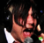 A pixelated image of a person with dark hair and glasses, wearing a white shirt, holding a microphone in one frame and a green object in another.