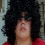 Pixelated image of a person with voluminous dark curly hair wearing sunglasses and red clothing, next to bold white text on black background.