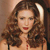 A pixelated close-up of a person with long brown hair, red lips, and a light-colored top against a warm-toned background.