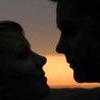 Two silhouetted figures face each other in profile, their lips meeting in a kiss against a gradient background of orange and gray.