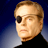 Pixelated static image of a man's face with an eyepatch, light brown hair, and black turtleneck against dark blue background.