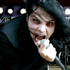 A person with dark hair and pale skin sings into a microphone, wearing black clothing against a stage background with lights.