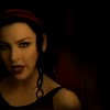 A static image of a person with dark hair and a red headband, wearing a black sleeveless top and a necklace with circular pendants, set against a dark background.