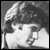 Black-and-white pixelated static icon showing a person's head and shoulders with dark hair, visible facial features including eyes and nose, and light-colored clothing against a gray background.