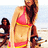 Two women in swimwear on a beach; one stands holding sunglasses, the other sits with arms crossed.