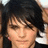 Pixelated close-up of a person with dark hair and light skin, wearing a dark top with a white collar, against a blurred background.