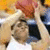 A person in a white tank top holds an orange basketball above their head with both hands, positioned against a blurred background of yellow and dark blue.