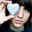 A person holds a light blue heart-shaped object over their right eye in three showing subtle facial expression changes
