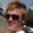 Pixelated static image of a person with light brown hair wearing dark sunglasses and a white shirt, set against a blurred outdoor background.