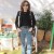 A pixelated image of a person standing outdoors wearing a dark long-sleeve top and light blue jeans, with a blurred background of greenery and structures.