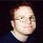 Pixelated static image of a person's face with light skin tone, dark hair, and glasses against a dark background.